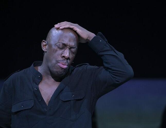 Images shows Giles Terera as Othello. He stands wearing all black, with his eyes closed, and his hand on his head a look of sorrow on his face. Photo by Myah Jeffers.