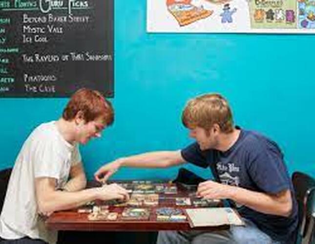 The image shows two young men sitting opposite each other playing a tabletop board game. They are both white, have short brown hair and are wearing tshirts.