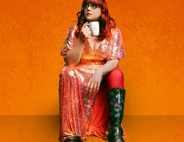Image shows comic Angela Barnes, with long red hair, sitting in an orange room. She's wearing a glittery orange dress, with one black boot showing, and she's drinking a cup of tea!