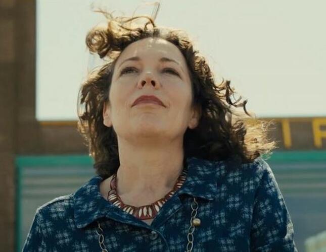 Image shows a woman, actor Olivia Colman, looking up and outward past the camera. The wind is blowing her dark curly hair away from her face. She is wearing a blue shirt and the background shows she is outside.
