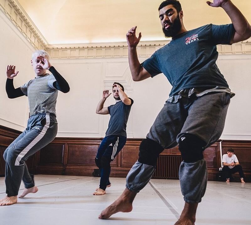 Image shows Three male dancers in a studio space are moving in unison with their arms raised and leg extended