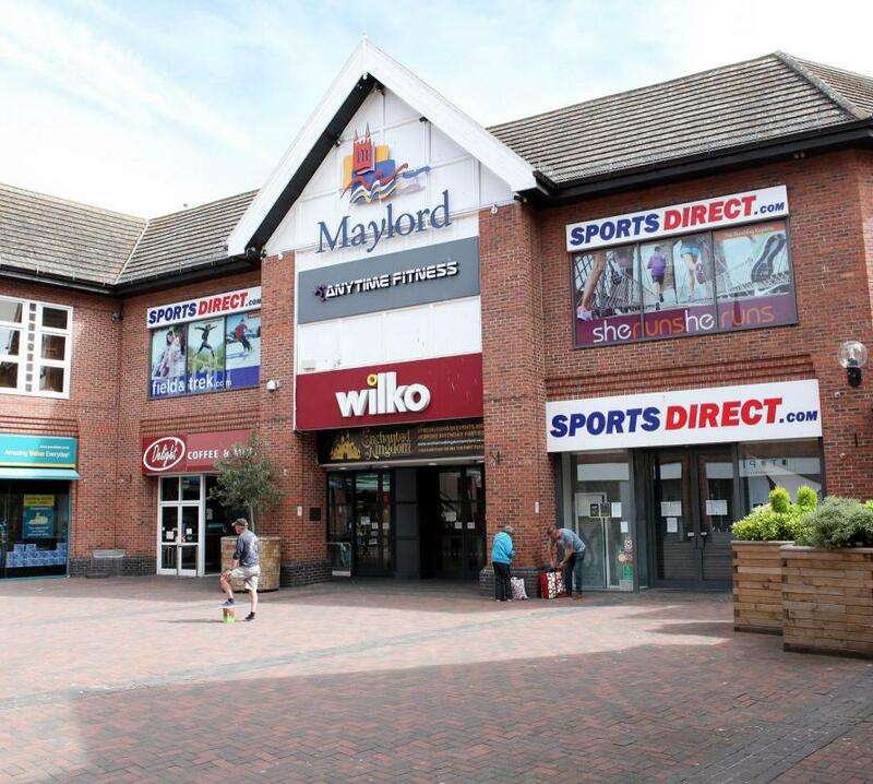 Image of a red brick and tile roofed shopping centre, with the name Maylords, in the roof apex. There are several shops inside, and three shoppers standing outside with bags.