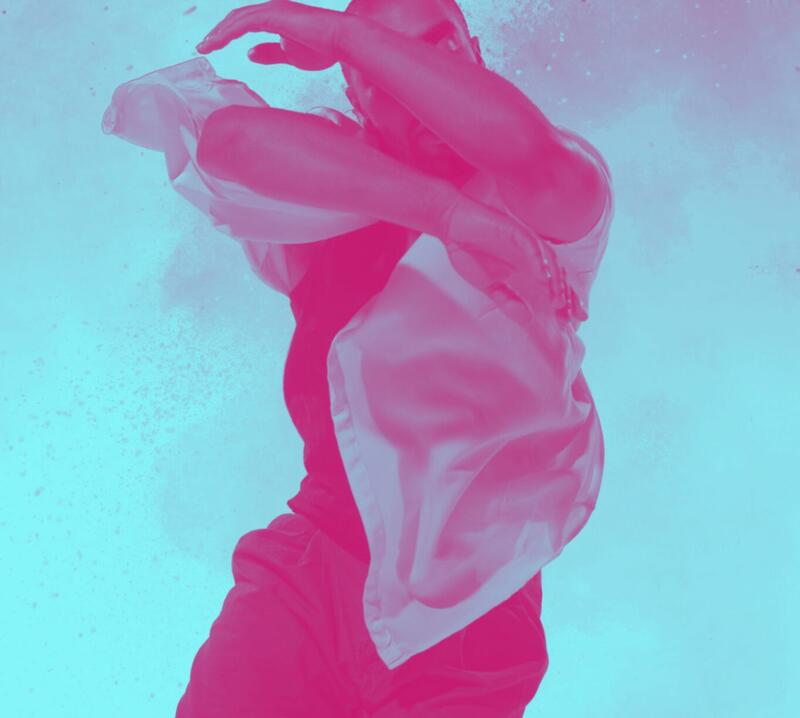 Images shows a male dancer moving fluidly, as if in water, his arms covering his face and a white shirt that is billowing. The colour of the image is manipulated to look bright pink on a blue background.