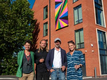 An image showing the geometric pattern artwork fixed to the side of the student accomodation building. Standing in front of the building in the foreground of the image is the artist Lothar Gotz, with art college principle Abigail Appleton, artist Daniel Pryde Jarman and Meadow Arts' Anne de Charmant. Photo by Oliver Cameron-Swan/Meadow Arts