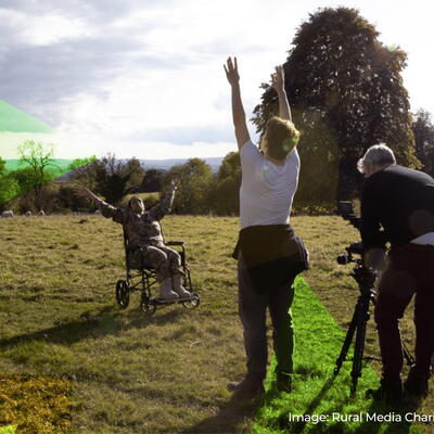 A person in a wheelchair raises their arms in a sunny meadow. Nearby, two people stand by a camera on a tripod. Trees and clouds in the background.