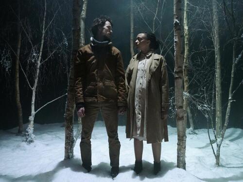 An image showing two people - a man and a pregnant woman - standing in a snowy forest. They are looking at each other. The sky is dark and the trees are bare.
