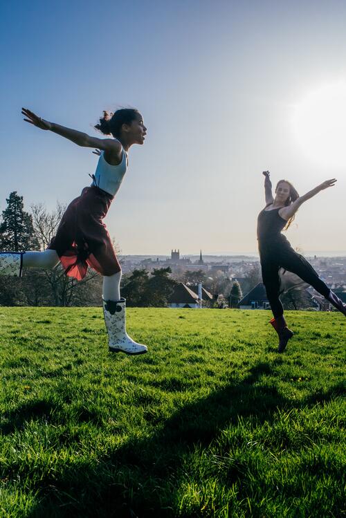 An image showing two dancers leaping in the air. They are outside on grass, and the sun is shining brightly.