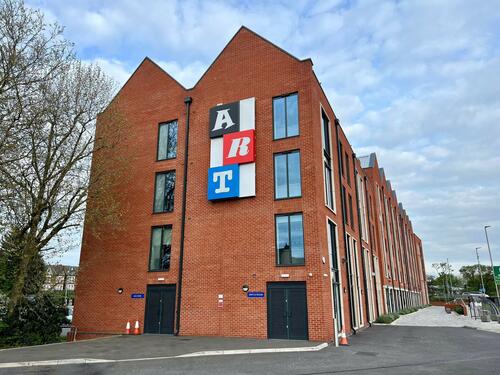 One of the new works at No 1 Station Approach by Matthew Cornford and John Beck. Image from Meadow Arts/HCA. Image showing the artwork attached to the side of a four storey red-brick building. The artwork is the word 'art' in stylised letters in black, red and blue