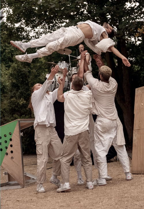 Image shows male dancers all wearing white. Three of them are throwing the fourth high over their heads into the air. His arms and legs are outstretched straight and long. The performance is taking place outdoors.