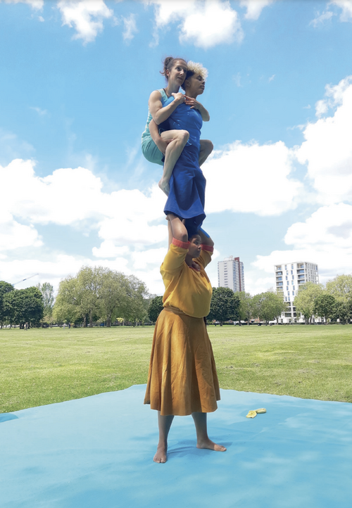 The image shows three women dancers who are acrobatically balanced on each others shoulders, so together they form a tall tower. They are wearing bright coloured dresses in yellow, blue and green. They are performing outside in a large urban park setting.