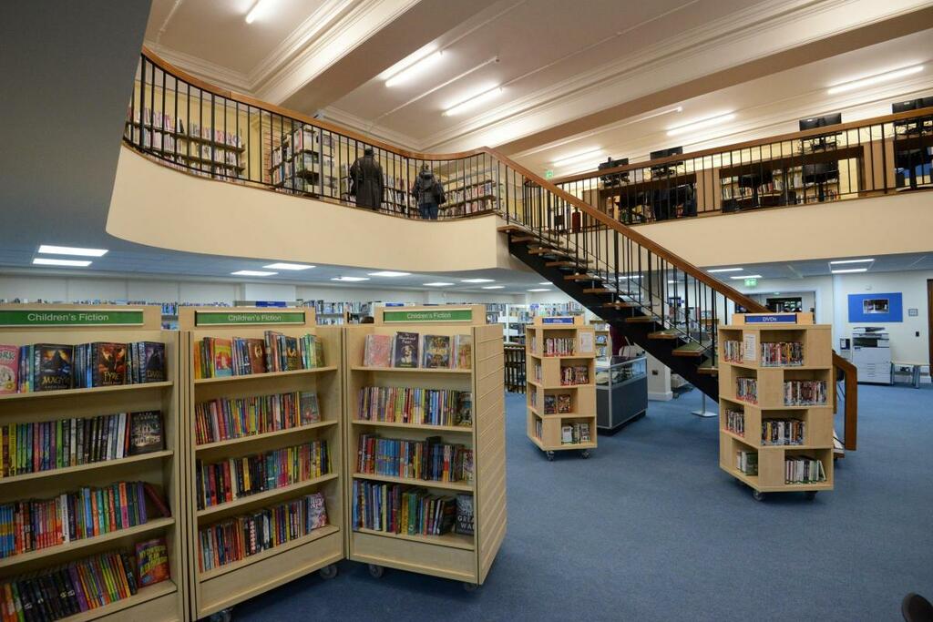 Image shows inside Hereford Library with an open plan layout and fully book cases in the foreground, and in the background a metal staircase that leads to an balcony floor.