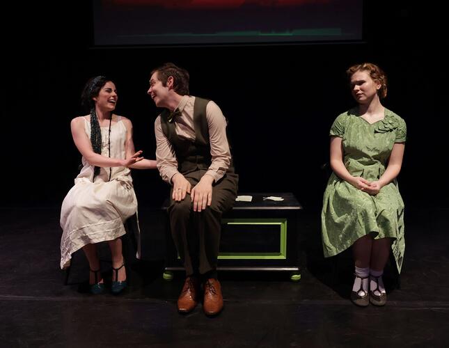 Image shows three people in 1920s clothing. A woman on thr right, sitting alone, wears a green dress and looks towards a man and a woman to her right. The man and woman are laughing together.