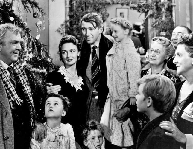 The image is a still from the black and white film It's a Wonderful Life and it shows a family gathered in a group with a Christmas tree in the background.