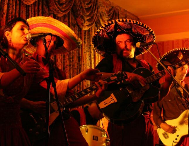 Image shows a live music on stage. The four musicians include a woman singing and three men playing guitars and wearing sombrero hats.