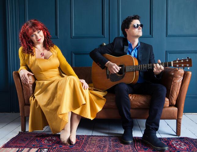 Image shows Roberts, a women with red hair sitting in a long yellow dress, next to Lakeman holding a guitar and wearing a dark suit and sunglasses