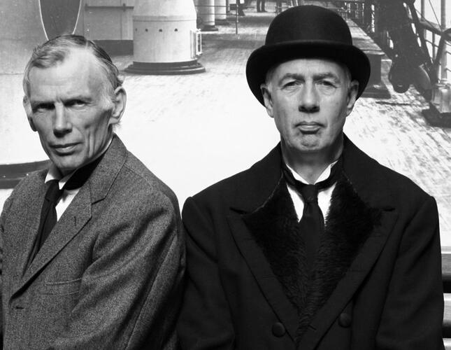 A black and white image of two actors - one man sits facing forward in a black bowler hat, the other man sits next to him in a grey suit.