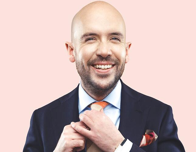 Image shows Tom Allen, a white man with bald head and stubble on this face, wearing a smart blue suit and smiling
