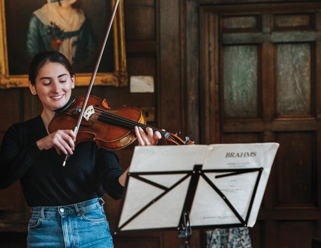 An image showing a smiling young woman playing violin in a luxourius looking panelled room. She is wearing a black shirt and demin jeans. 