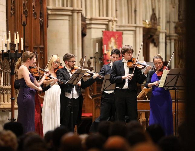 An image show the classical music ensemble members standing on a stage in a church setting. They are wearing dinner suits and dresses.