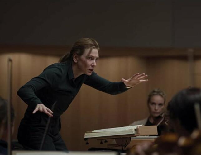 Image is a still from the film which shows Cate Blanchett in the role of a conductor. She is wearing all black and has her hands spread wide as she conducts orchestra members who are not in the image.