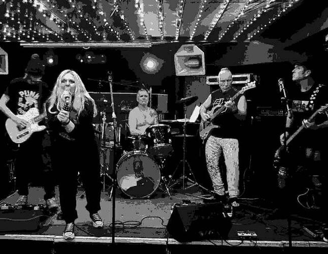 Image is black and white and shows a punk band playing live on stage. The lead singer is a woman with long blonde hair.