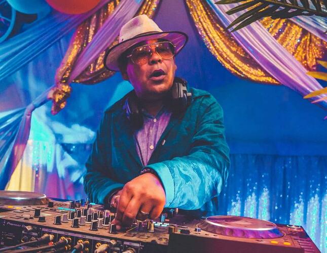Image shows DJ Craig Charles using a set of decks. He's wearing a hat and sunglasses and blue jacket. The background is the inside of a brightly coloured tent.