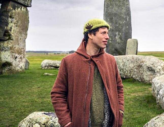 Image shows folk singer Sam Lee, wearing a yellow hat and brown jacket, looking off to the right. He is standing among ancient standing stones.