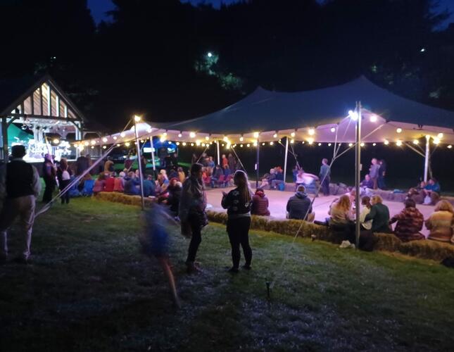 People sitting under fairy light festooned marquee at night, next to a bandstand
