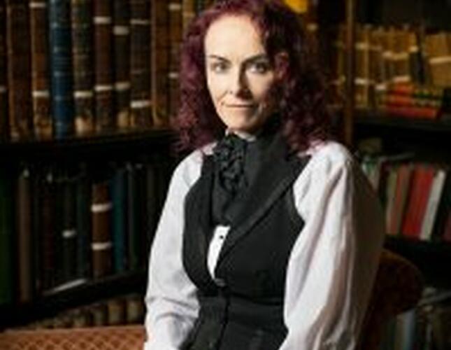 Rosie Garland, with curly hair, seated in a dark library wearing a white shirt with ruffled sleeves and a black waistcoat. 