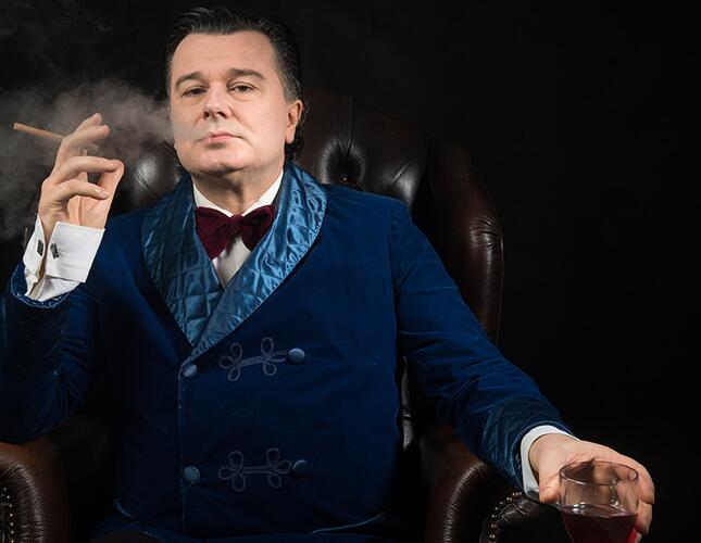 An actor looking straight at the camera wearing a blue suit smoking a cigarette