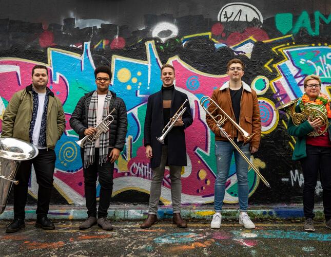 a group of young people holding brass instruments standing in front of a wall with graffiti